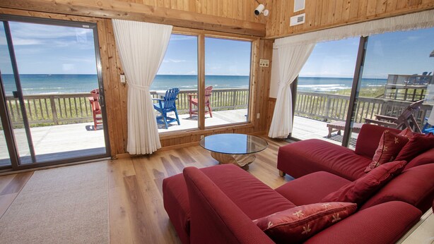 Een houten interieur met een rode bank, een glazen tafel en uitzicht op het strand door grote ramen.