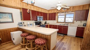 Een keuken met houten kastjes, een magnetron, een fornuis en een koelkast. Er staan drie krukken met rode kussens.