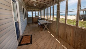 En overdækket træterrasse med bænk og bord, med udsigt over en strand.