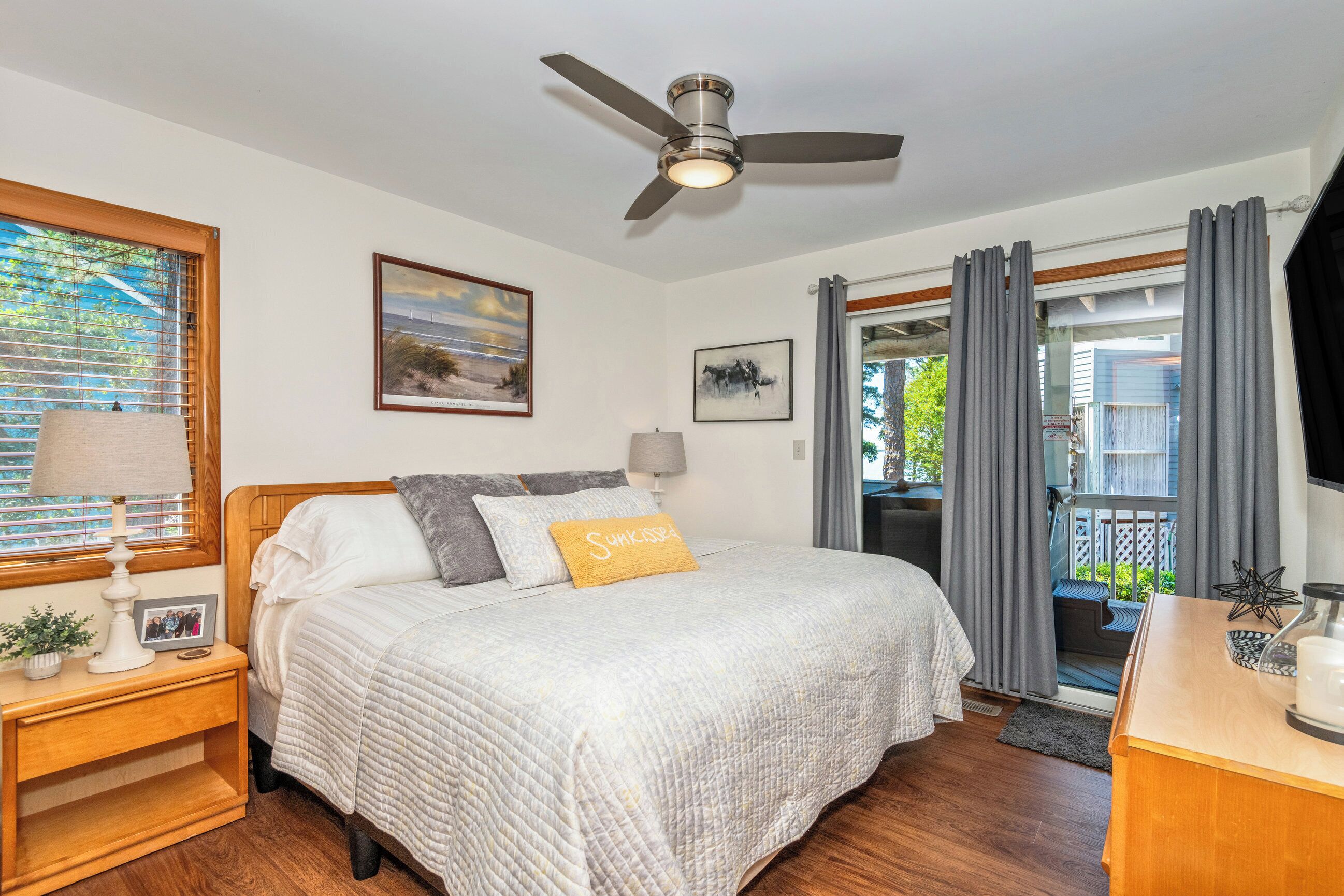 A bedroom with a bed, a nightstand, a ceiling fan, a window with blinds, and a view of the outside.