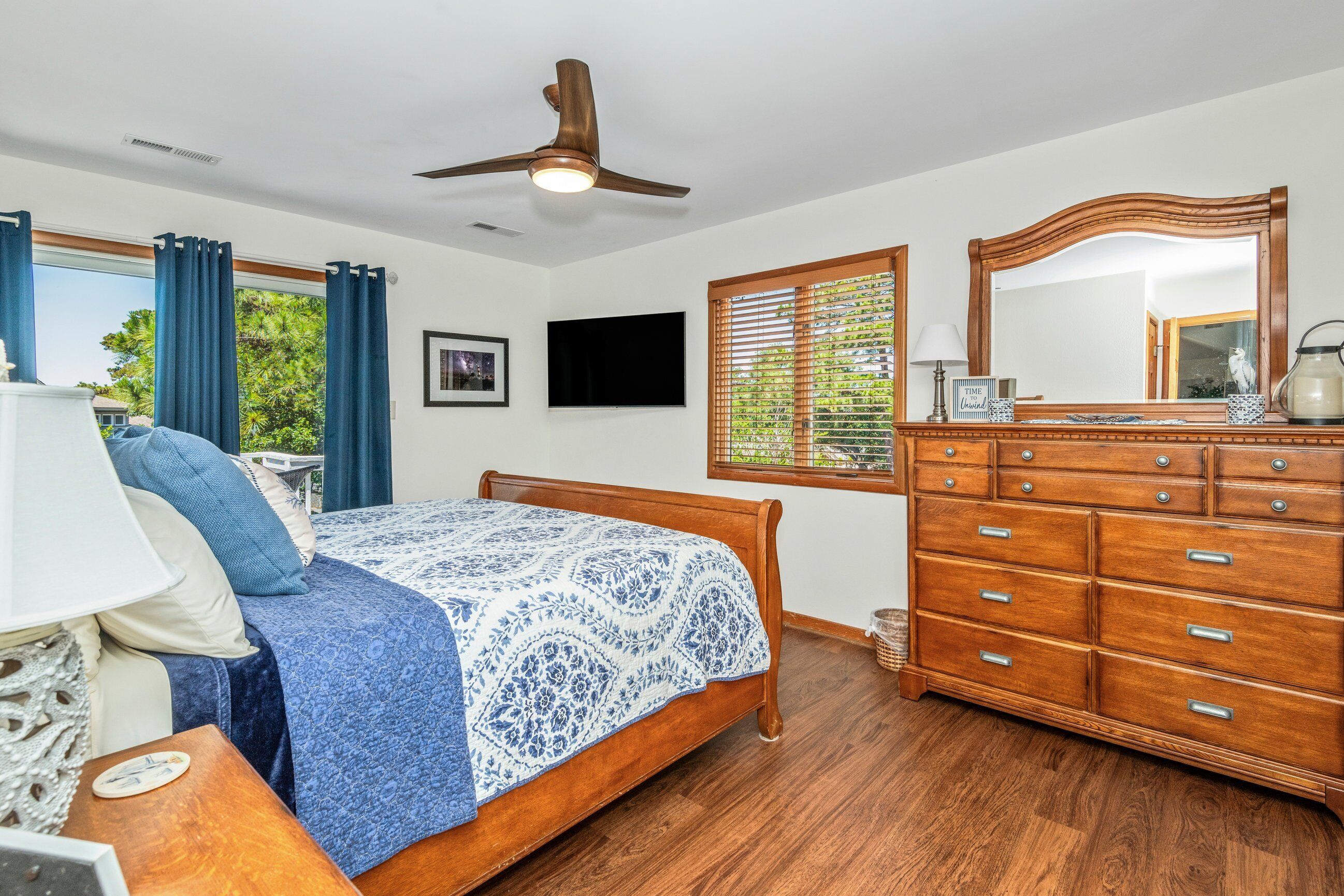 A bedroom with a bed, wooden dresser, ceiling fan, TV, window with blinds, and a framed picture on the wall.