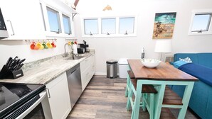 A modern kitchen with white cabinets, a stove, and a sink. There is a dining table with chairs and a blue sofa in the living area. - Semi-Oceanfront, Pets Welcome, Wild Horses, Ocean Views! 4x1678, Turtle Trax (Corolla)