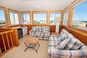 Una habitación acogedora con kitchenette, un conjunto de sofás y ventanas amplias con vistas al mar y casas vecinas.