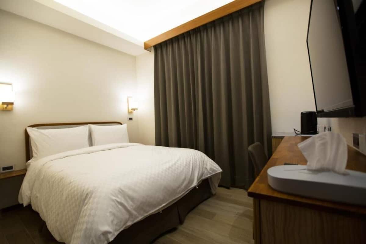 A hotel room with a bed, a desk with a chair, a television, and a window with curtains.