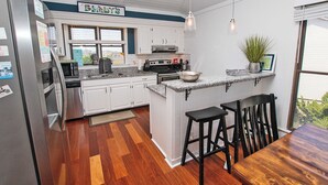 A modern kitchen with white cabinets, a stainless steel refrigerator, a wooden dining table, and a bar area with stools. - Oceanside, Close to Beach Access, Community Pool, KHSDD1, Sea Dunes D1 (Kitty Hawk)