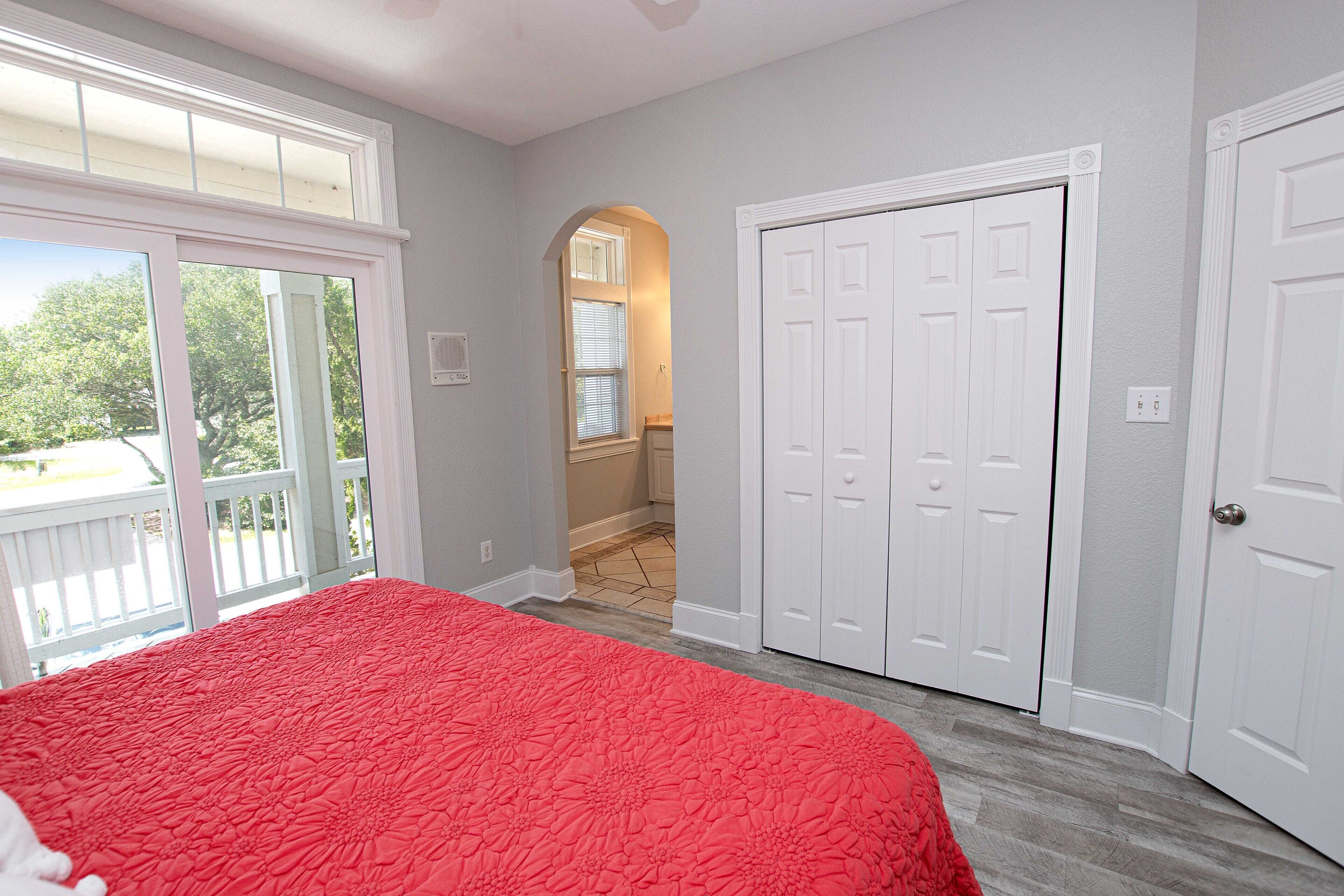 Une chambre à coucher avec une couvre-lit rouge, des portes blanches et une vue sur l’extérieur par une fenêtre.