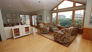 A spacious living room with a high ceiling, large windows offering a view of houses, and a white entertainment unit with storage baskets.