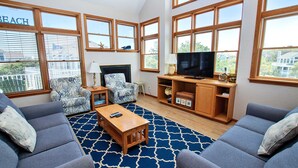 A spacious living room with large windows, a fireplace, a television, and a blue patterned rug. - Oceanside, Private Pool, Volleyball, Screened porch, PI61, Without a Porpoise (Corolla)
