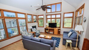 A spacious living room with a large grey sofa, a television, and a ceiling fan. - Oceanside, Private Pool, Volleyball, Screened porch, PI61, Without a Porpoise (Corolla)