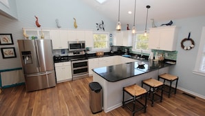 A modern kitchen with white cabinets, stainless steel appliances, a large island with bar stools, and decorative wall art. - Oceanside, Private Pool, Walking distance to town of Duck, Foosball, T3, Bella Vita (Duck)