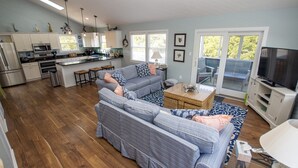 A spacious living area with a grey striped sofa, a small coffee table, and a TV stand. A kitchen with white cabinets and a dining area with a table and chairs is visible. - Oceanside, Private Pool, Walking distance to town of Duck, Foosball, T3, Bella Vita (Duck)