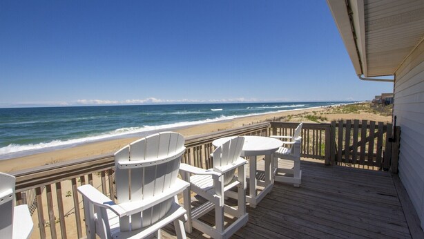 A wooden deck with white Adirondack chairs and a small table overlooking a beach with waves.