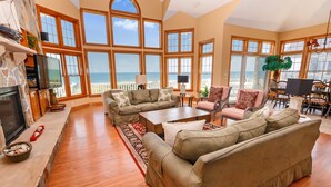 A spacious living room with a large window overlooking the ocean, a fireplace, and a dining area with outdoor seating. - OCEANFRONT, Ocean views, beach access, Pool, PI139, Pine Haven (Corolla)