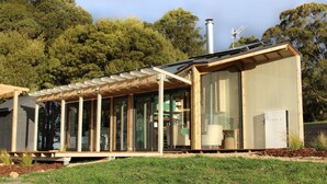 Une maison moderne dotée de grandes baies vitrées, d’une terrasse en bois et d  ’une borne de recharge pour véhicules Tesla.