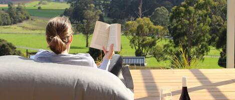 Una persona leyendo un libro en una cubierta de madera con vistas a un paisaje verde.