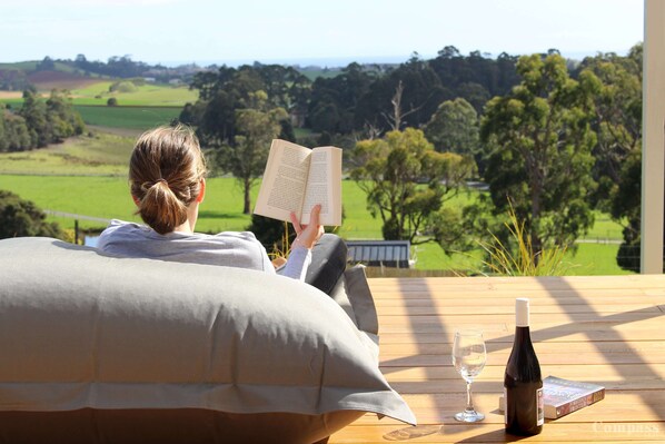 A person reading a book on a wooden deck with a view of a green landscape.