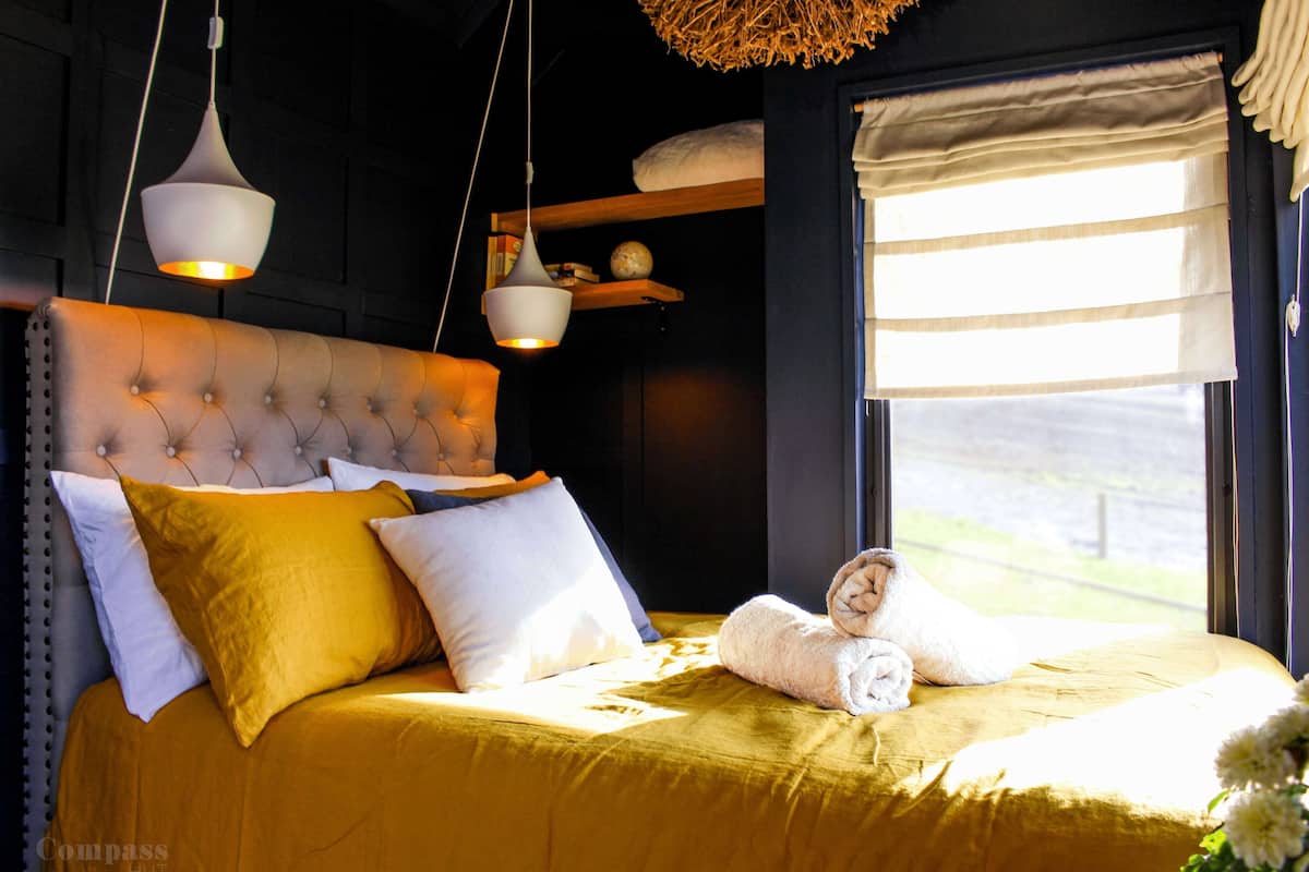 Un lit doté d’une tête de lit capitonnée, d’une couvre-lit jaune et d’oreillers blancs et moutarde. Des luminaires suspendus sont placés au-dessus du lit, et une fenêtre est agrémentée d’une lambrequin à rayures.