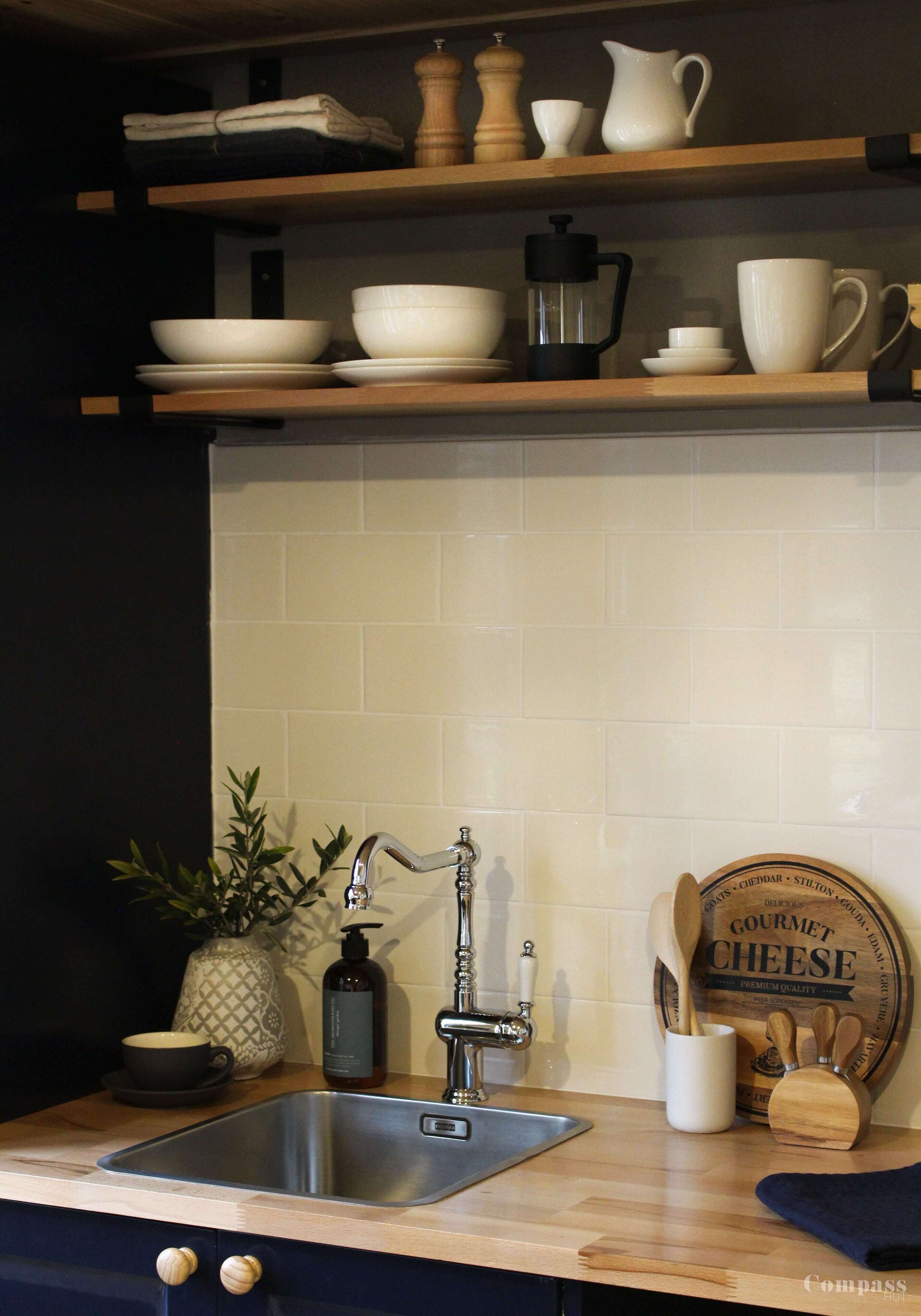 A kitchen with a stainless steel sink, wooden shelves above holding bowls and a coffee pot, a potted plant, and a cheese board.