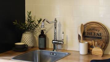 A kitchen with a stainless steel sink, wooden shelves above holding bowls and a coffee pot, a potted plant, and a cheese board.