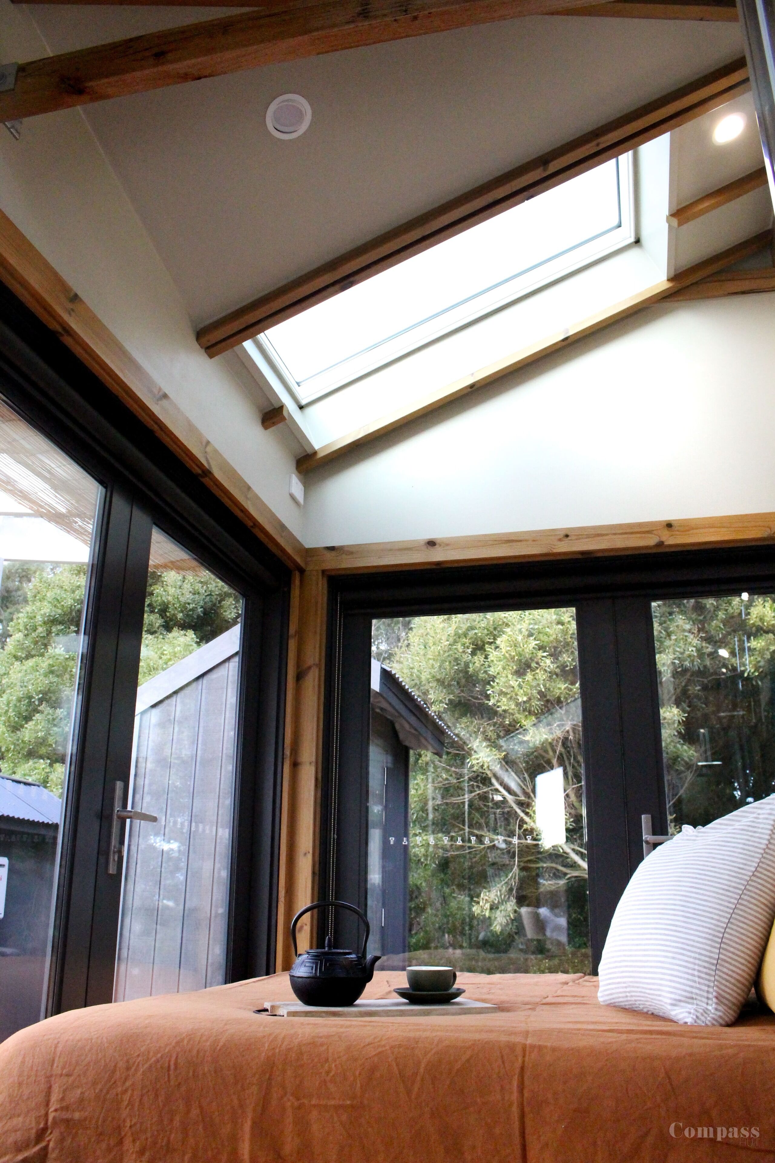 A bedroom with a skylight, a bed with a brown bedspread, a black kettle on a small table, and a large window with a view of trees and a house.