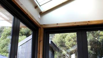 A bedroom with a skylight, a bed with a brown bedspread, a black kettle on a small table, and a large window with a view of trees and a house.