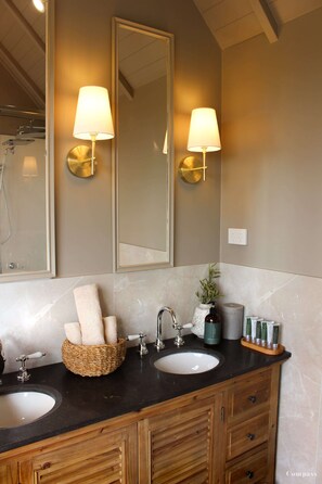 A bathroom with two sinks, a mirror, wall-mounted lamps, a towel rack, and a wicker basket.