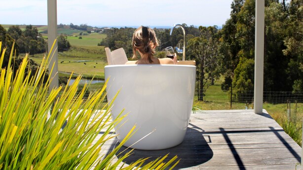 Une femme se détend dans une baignoire blanche sur une terrasse en bois, avec une vue panoramique sur un paysage verdoyant et une étendue d’eau en arrière-plan.