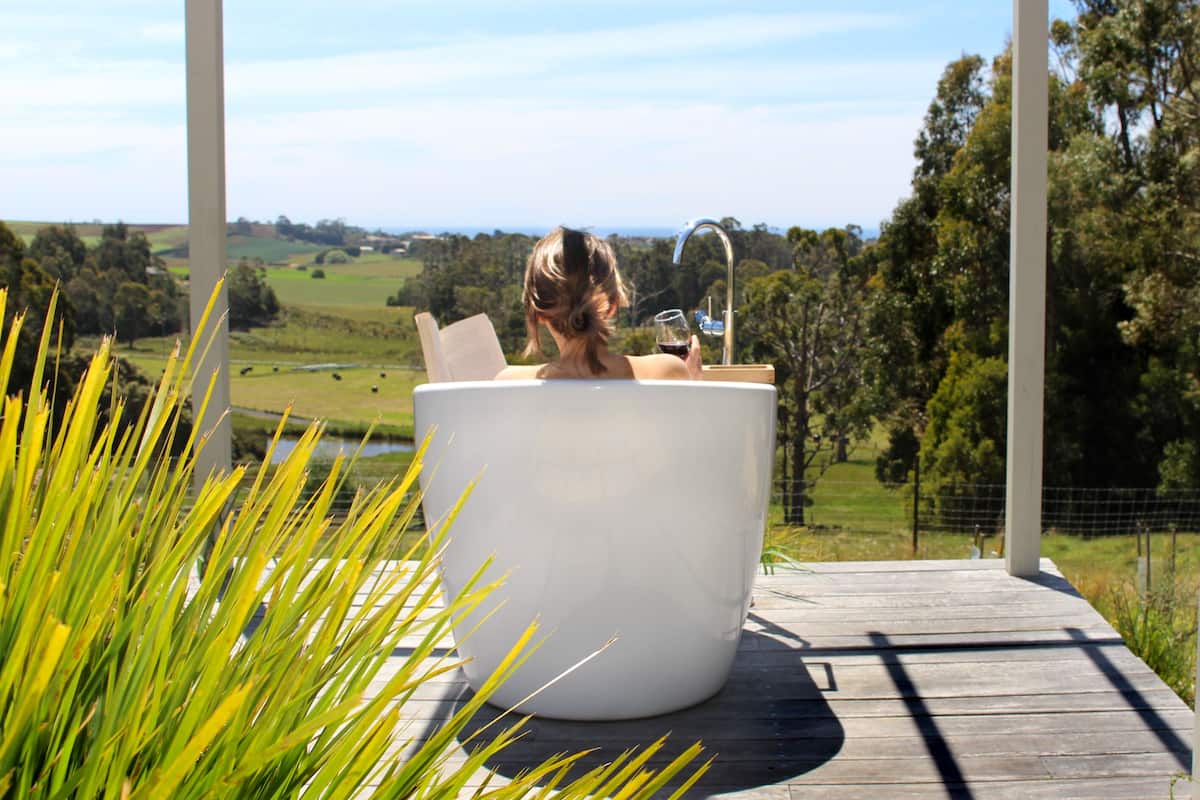 Une femme se détend dans une baignoire blanche sur une terrasse en bois, avec une vue panoramique sur un paysage verdoyant et une étendue d’eau en arrière-plan.