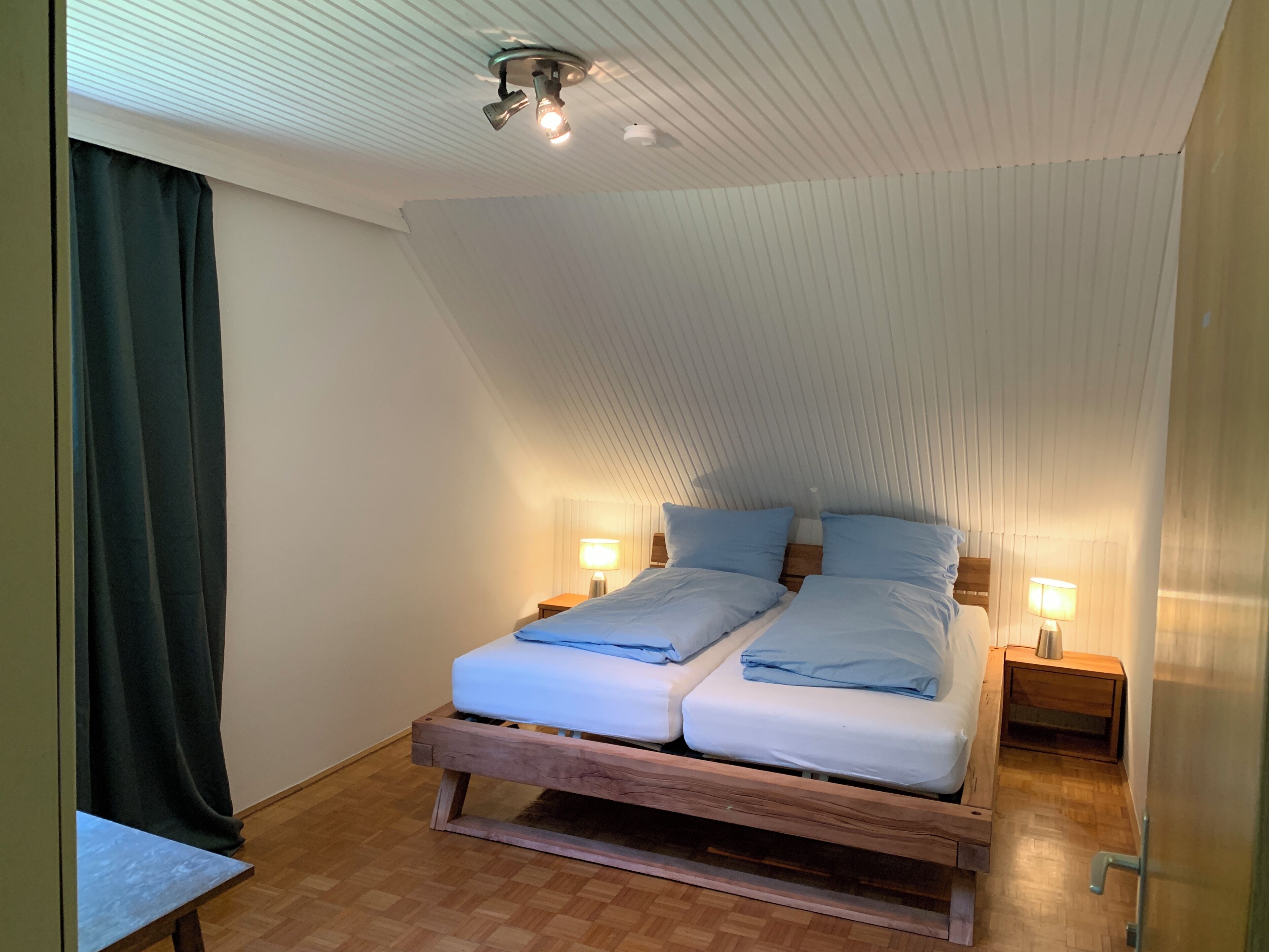 A room with a sloped ceiling, two beds with blue bedding, wooden headboards, bedside lamps, and a wooden bench.
