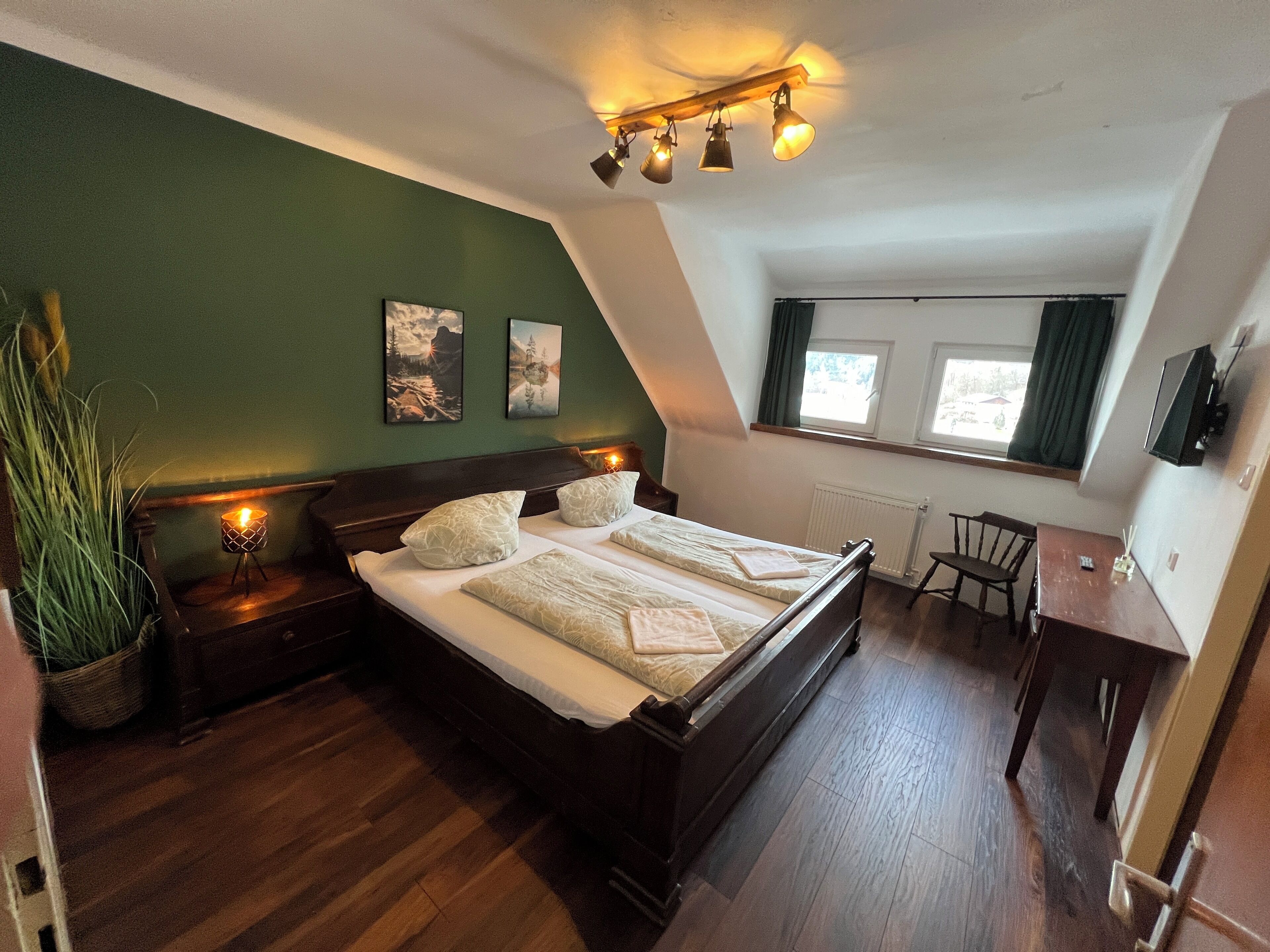A bedroom with a wooden bed, a small desk, a chair, a radiator, and a window with curtains.