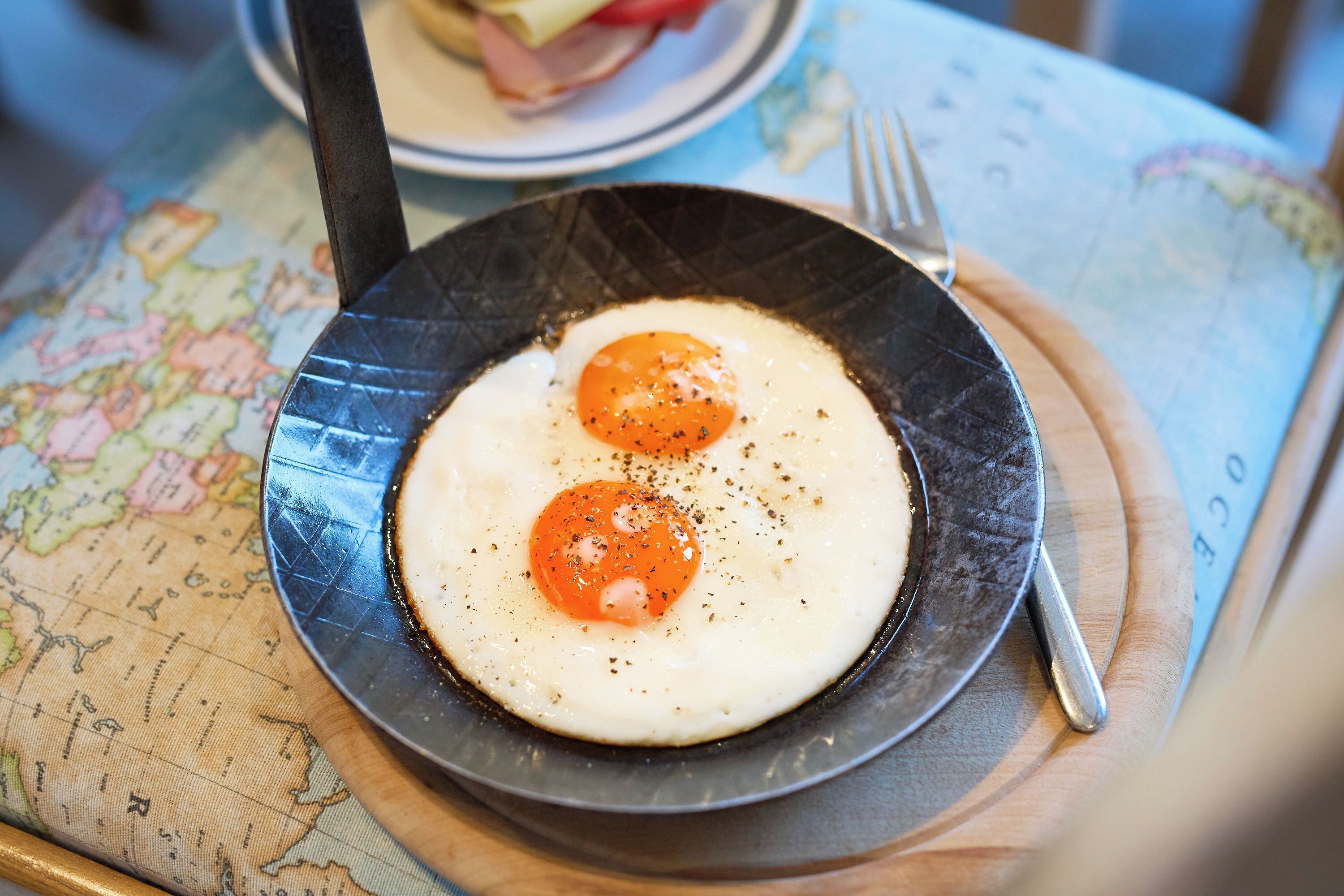 A black skillet with two sunny-side-up eggs on a wooden board, placed on a table with a map.