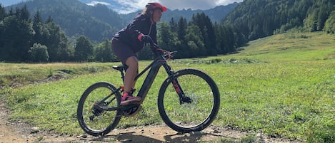 Una persona montando una bicicleta de montaña sobre un sendero de tierra en un campo cubierto de hierba, con montañas al fondo.