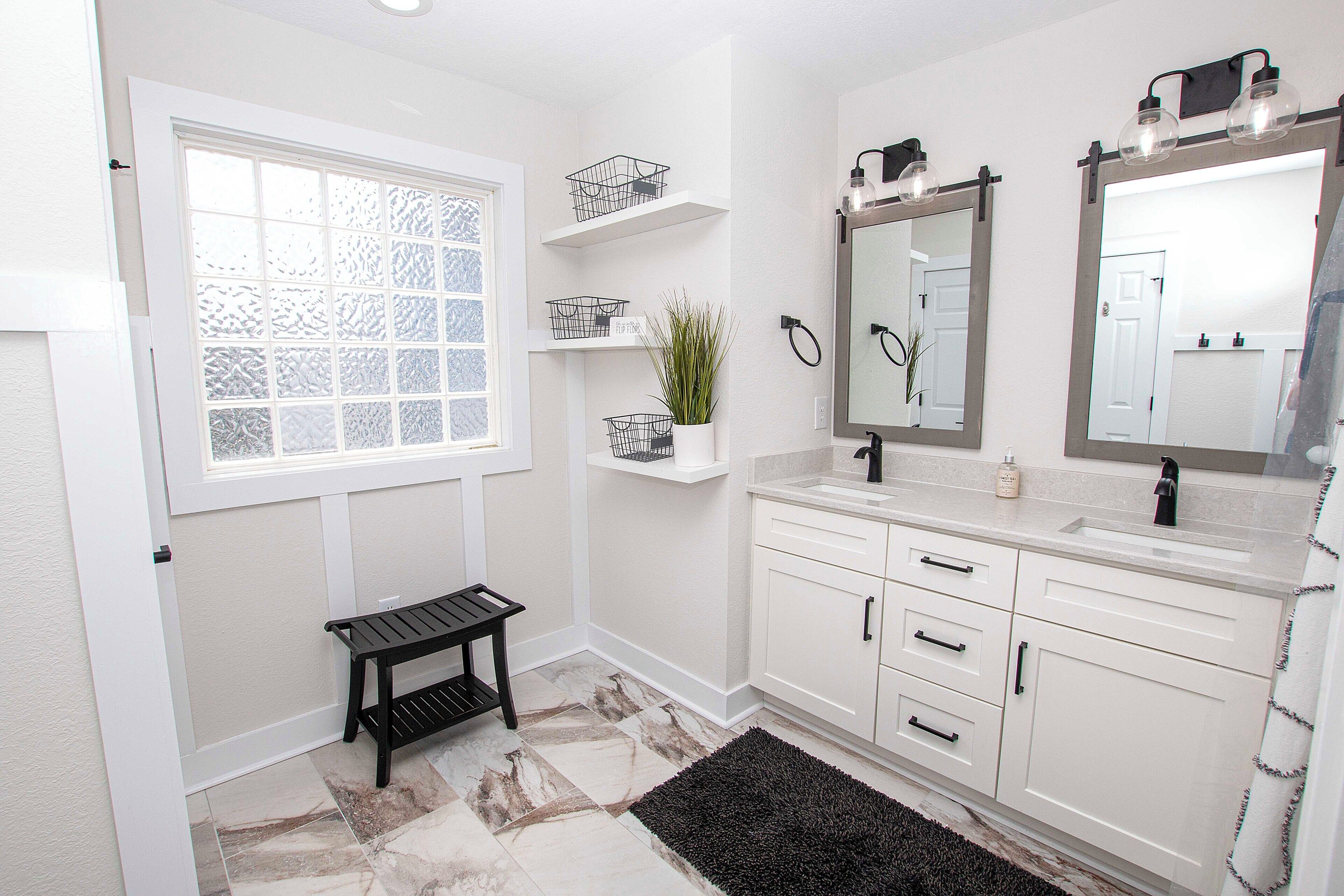 A bathroom with a white vanity, a black bench, a window with frosted glass, and a mirror with lights above it.