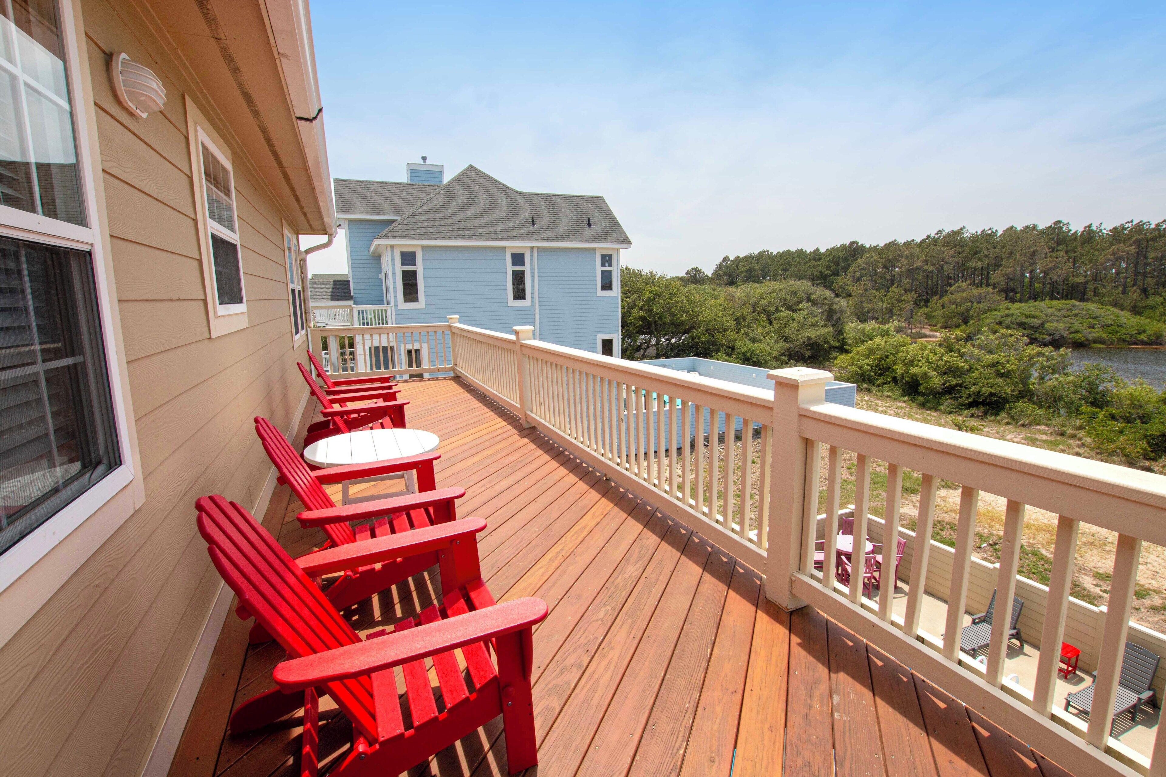 A wooden deck with red Adirondack chairs and a white railing overlooking a body of water and forested area.