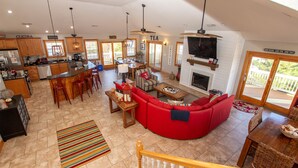 A spacious living area with a red sofa, a dining table, and a kitchen area with wooden cabinets and a TV mounted above the fireplace. - Oceanside, 8 BRs, Private Pool and Hot Tub, Pool Table, PI109, Sweet TEA (Corolla)