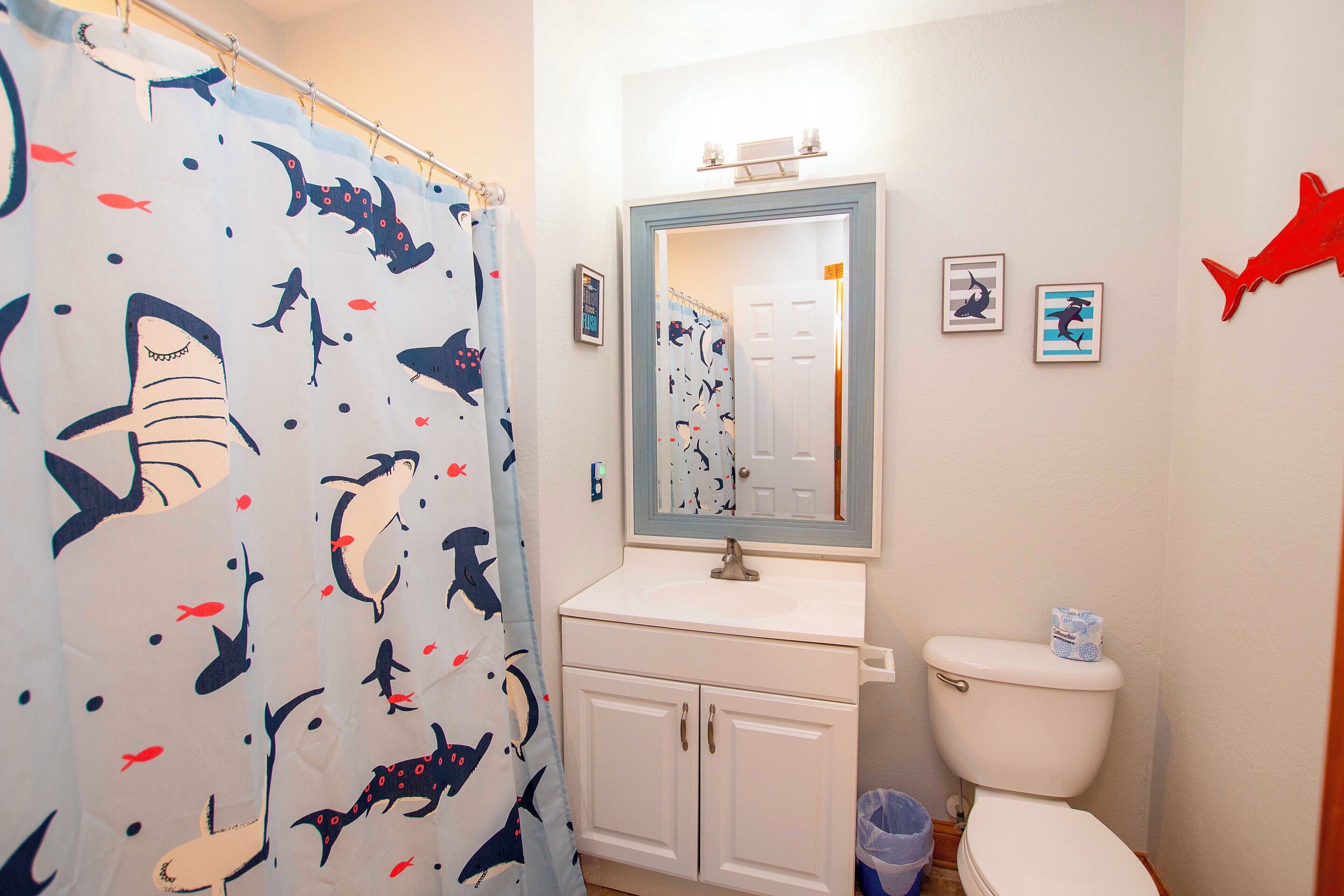 A bathroom with a shark-themed shower curtain, a white sink, a mirror above the sink, and a toilet. There are framed pictures on the wall and a red shark decoration.