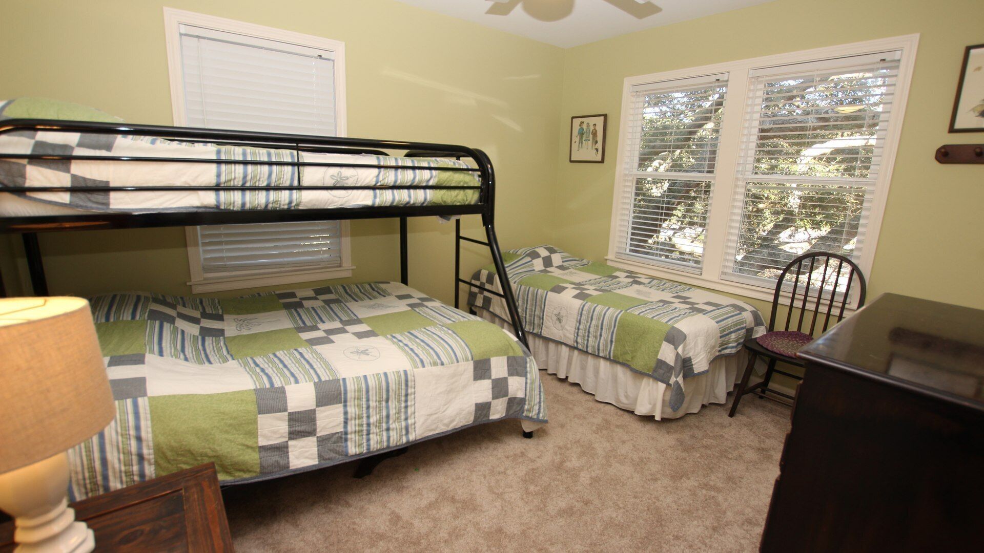 A dormitory room with bunk beds, a desk, and a chair. There are two windows with blinds, a lamp, and a framed picture on the wall.