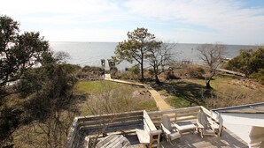 En træterrasse med Adirondack-stole med udsigt over vand, omgivet af træer og en græsplæne.