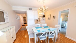 A dining area with a white table and chairs, a chandelier, and a kitchen in the background. - Oceanfront, Community Pool and Beach Accesses, Ocean Views, C326D, Our Point Of View (Duck)