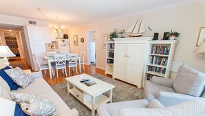 A living room with a white sofa, a coffee table, and a bookshelf. - Oceanfront, Community Pool and Beach Accesses, Ocean Views, C326D, Our Point Of View (Duck)