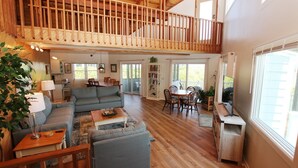 A spacious living area with a high ceiling, wooden beams, a dining area with a table and chairs, and a balcony with a railing. - Oceanside, Private Pool, Screened Porch, Close to Beach Access, OS3H, Brigadune (Corolla)