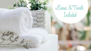 A neatly folded white towel with a woven basket and dried lavender in the background. Text indicates linens and towels are included.