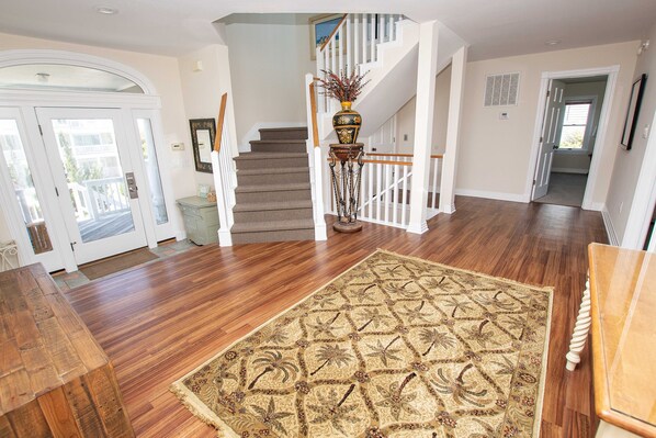 A spacious foyer with a wooden floor, a large patterned rug, a staircase with white railings, and a door leading outside. - OCEANFRONT, Ocean Views, ELEV, Pool, Pool Table, Priv Beach Access, PI187, Right On The Beach (Corolla)