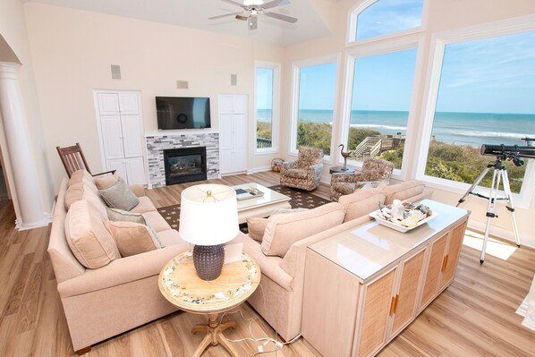 A spacious living room with a large window overlooking the ocean, a fireplace, and a television mounted on the wall. - OCEANFRONT, Ocean Views, ELEV, Pool, Pool Table, Priv Beach Access, PI187, Right On The Beach (Corolla)