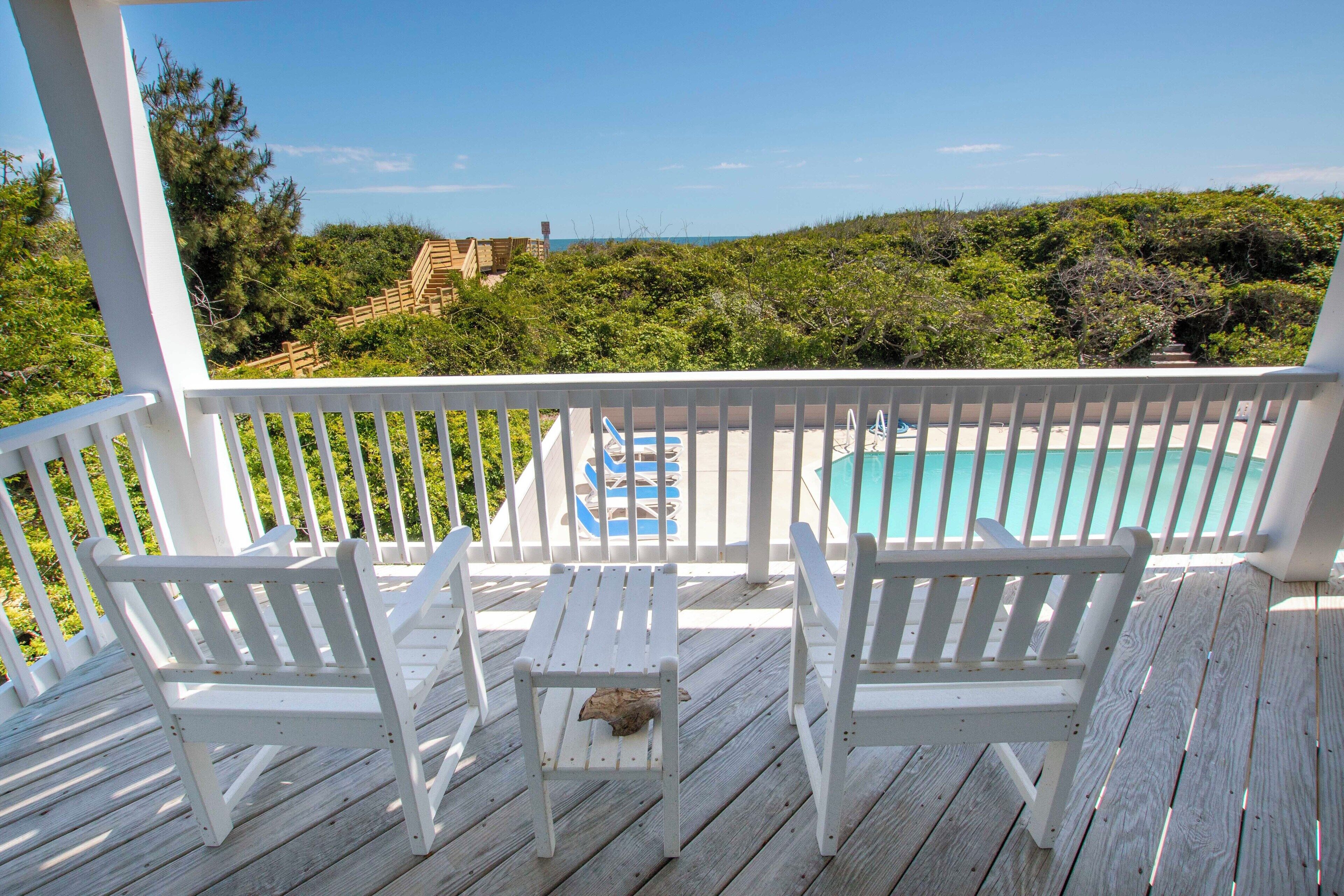 A wooden deck with white chairs and a small table, overlooking a swimming pool and a view of the ocean.