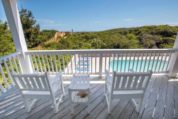 Una terraza de madera con sillas blancas y una pequeña mesa, con vista a una piscina y abundante follaje.