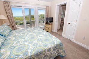 A bedroom with a bed, a television on a wooden cabinet, a balcony with a view of the landscape, and a bathroom visible through an open door.