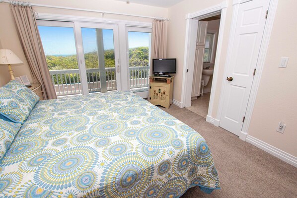 Une chambre à coucher avec un lit, un téléviseur posé sur un meuble en bois, un balcon offrant une vue sur le paysage, et une salle de bain visible à travers une porte ouverte.