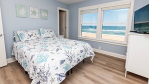Un dormitorio con cama, ventilador de techo, ventana con vista a la playa y decoración en la pared.
