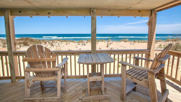 Una terraza de madera con dos sillas Adirondack y una pequeña mesa, con vista a una playa con dunas de arena y el océano.
